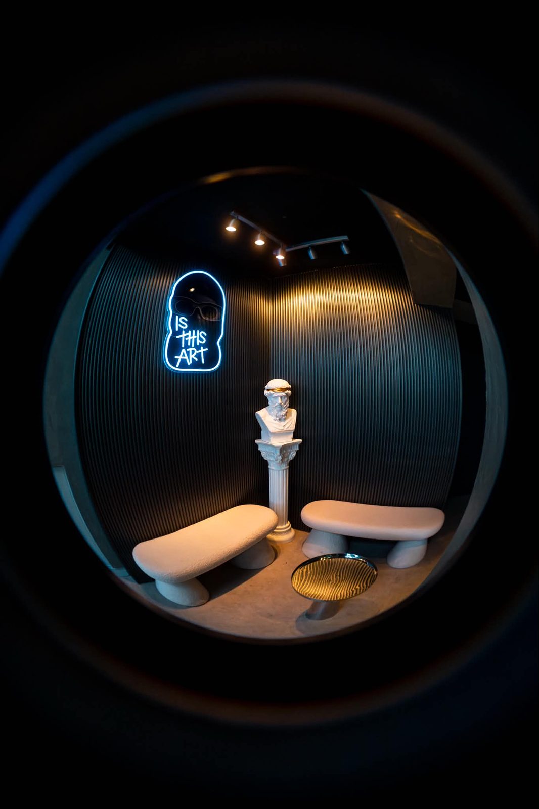 The waiting corner at Culture Box: a cyan neon sign reading IS THIS ART above a marble bust wearing a gold laurel, between two bouclé benches.