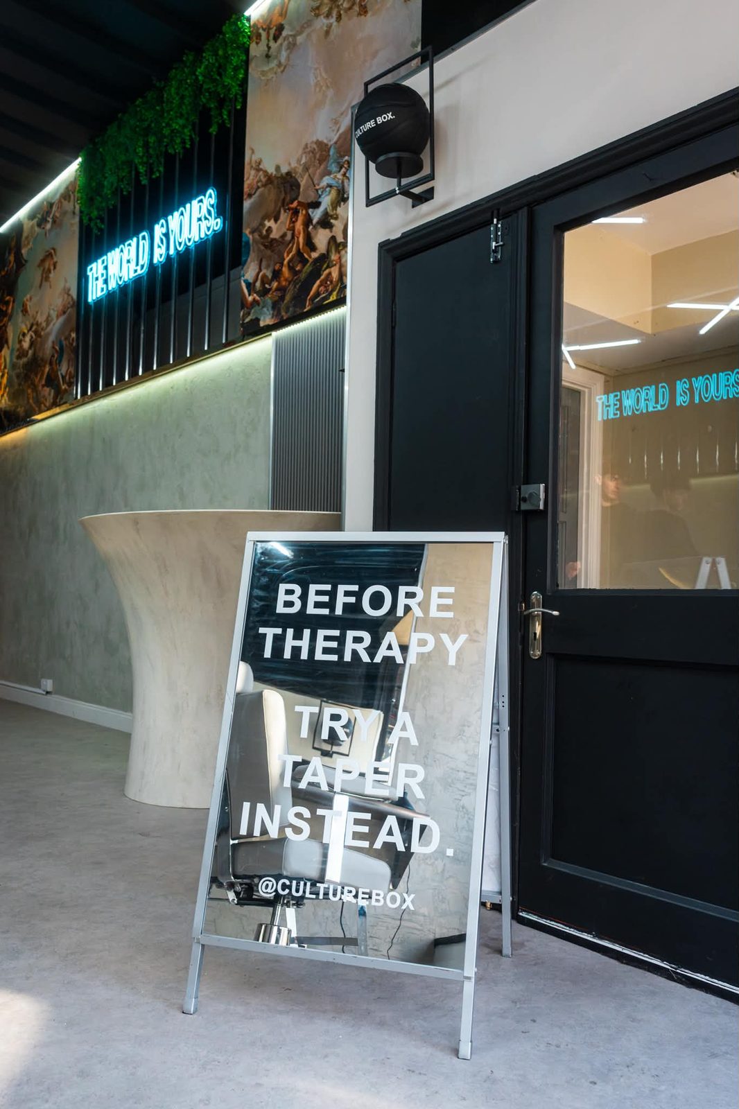 The entrance at Culture Box: a mirrored A-board reading BEFORE THERAPY TRY A TAPER INSTEAD, beside the sculptural reception desk and the neon.